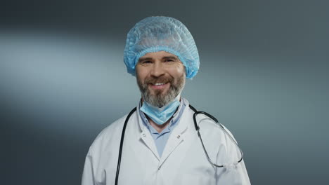 Portrait-shot-of-the-Caucasian-handsome-man-doctor-in-blue-hat-taking-off-mask-from-his-mouth-and-smiling-cheerfully-to-the-camera.